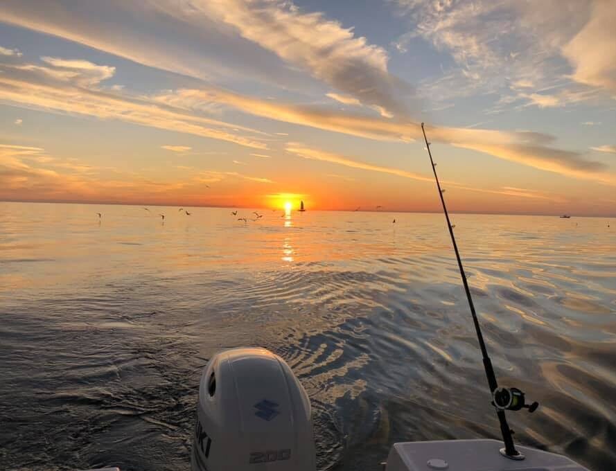 A fishing pole hangs of the stern of a boat in front of a sunset.