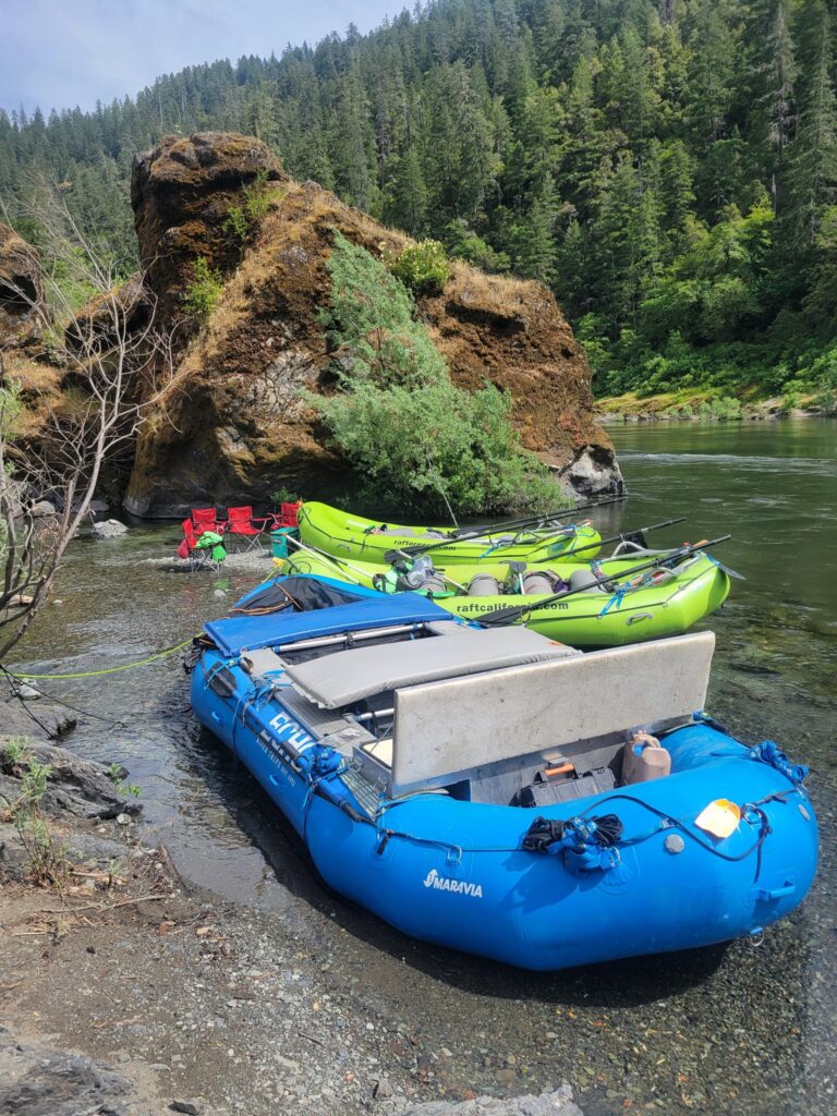 One blue raft and two green rafts beached on the side of a river.
