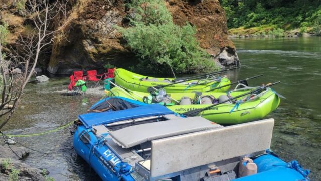 One blue raft and two green rafts beached on the side of a river.