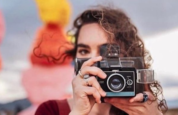 A girl holding a camera with a rainbow sculpture in the background