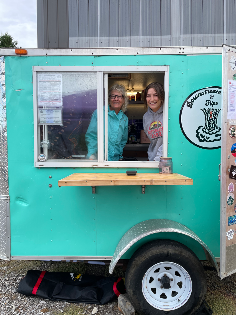 Two Ladies poking their heads out of a tiny blue food truck trailer.
